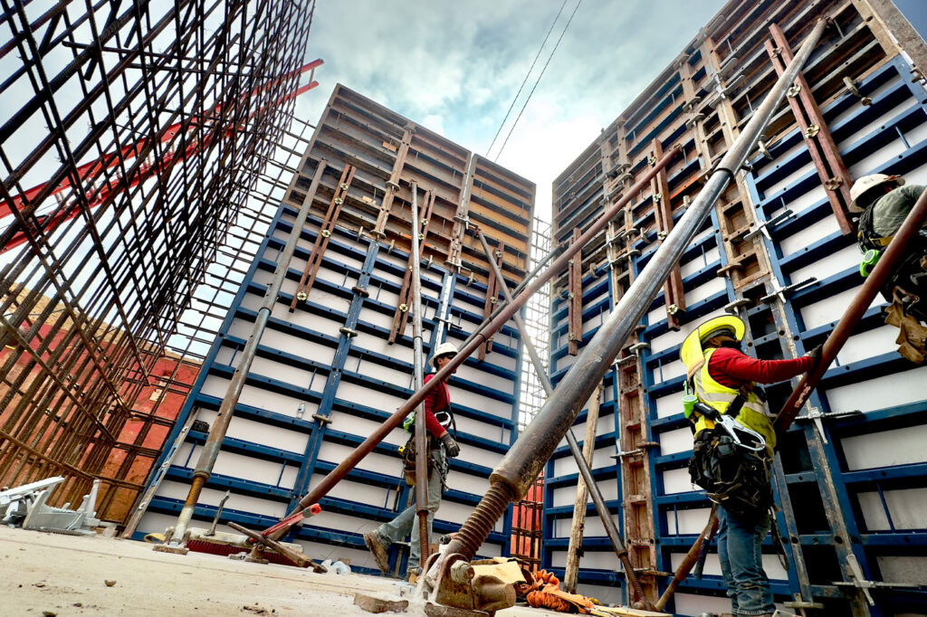 Construction workers in safety gear work on a building site surrounded by tall metal scaffolding and large blue and white concrete forms under a cloudy sky.