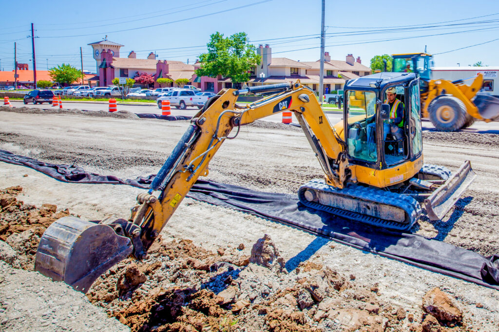 A construction worker operates a small yellow excavator, digging up dirt on a road under construction. Orange barrels line the site, and vehicles and buildings are visible in the background on a sunny day.