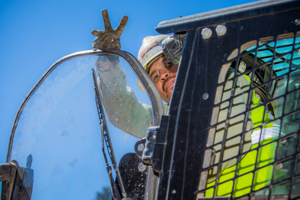 A construction worker in a helmet and safety vest raises a gloved hand, making a peace sign while sitting inside heavy machinery, with a bright blue sky in the background.