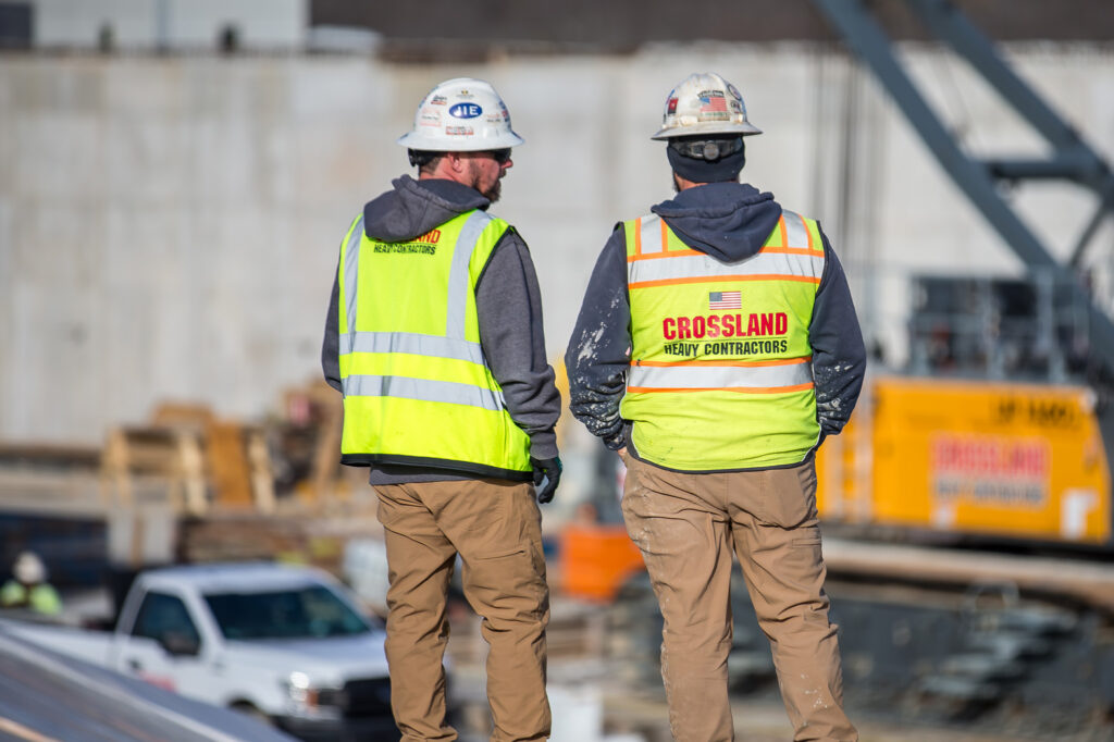 Two construction workers in yellow safety vests and hard hats stand at a job site, facing away. One vest reads “CROSSLAND HEAVY CONTRACTORS.” Construction equipment and vehicles are visible in the background.