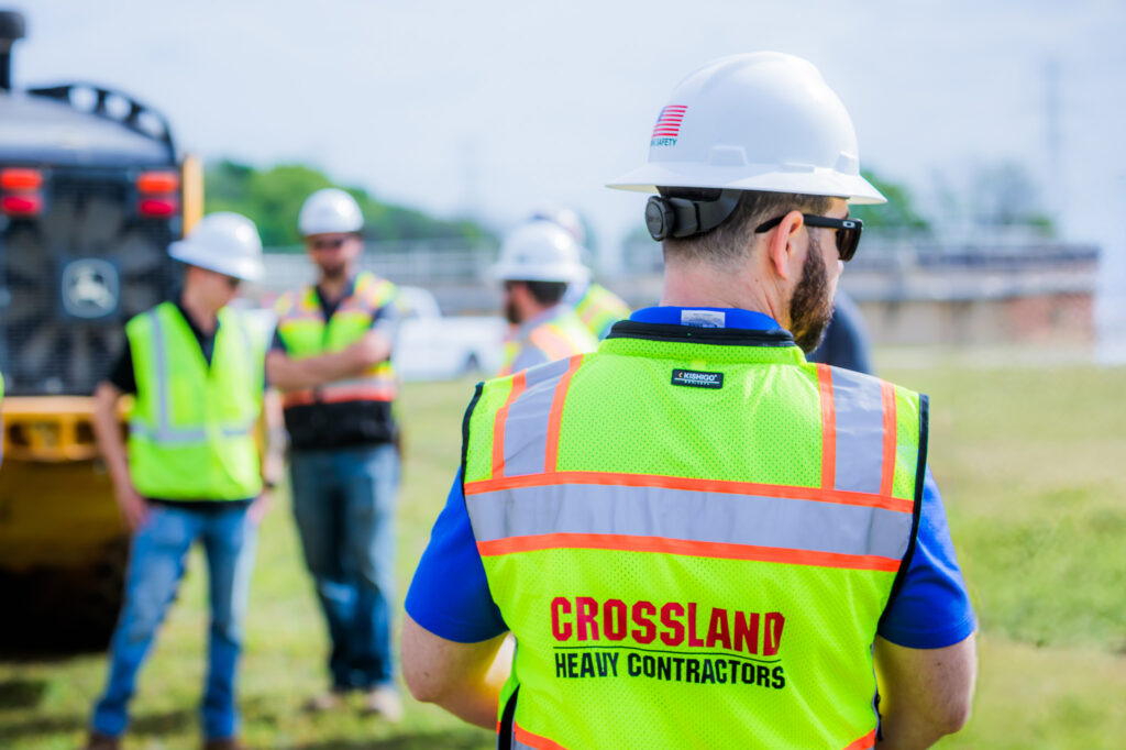 A construction worker wearing a white helmet and a safety vest labeled “Crossland Heavy Contractors” stands outdoors, with three other workers in safety gear talking in the blurred background.