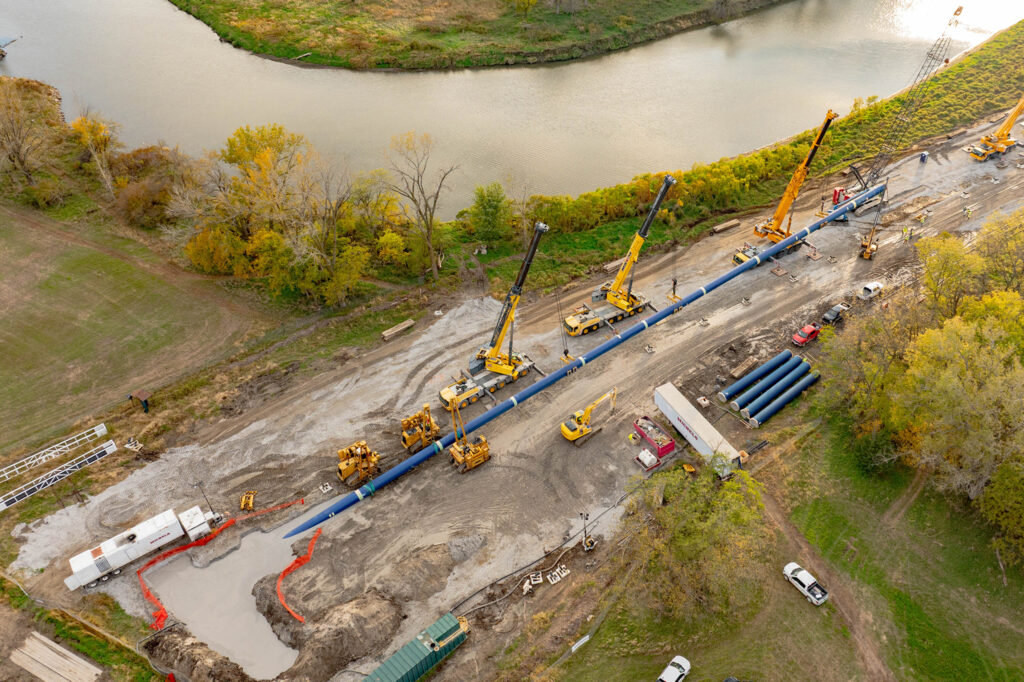 Aerial view of a construction site near a river with cranes, vehicles, and workers installing large blue pipes along a cleared, dirt path surrounded by trees and grassy areas.