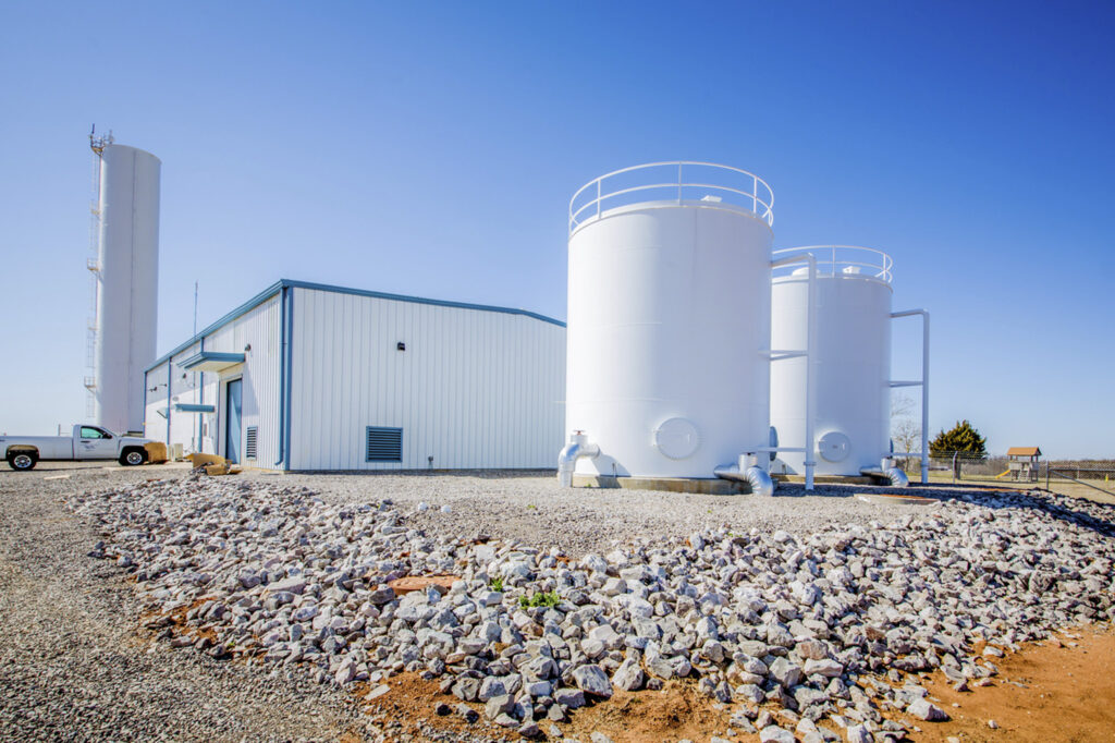 Large white industrial storage tanks and a metal building sit on a gravel lot under a clear blue sky; a white pickup truck is parked nearby.