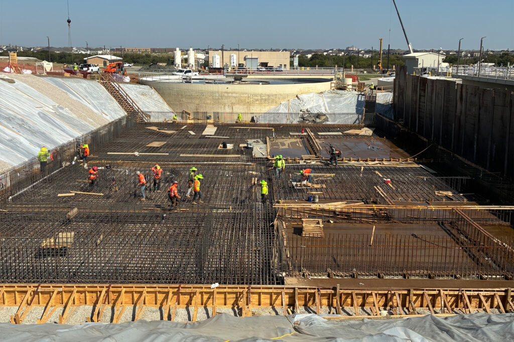 Construction workers in safety gear work on a large building foundation, surrounded by rebar, wood forms, and construction materials, with industrial buildings and equipment visible in the background.