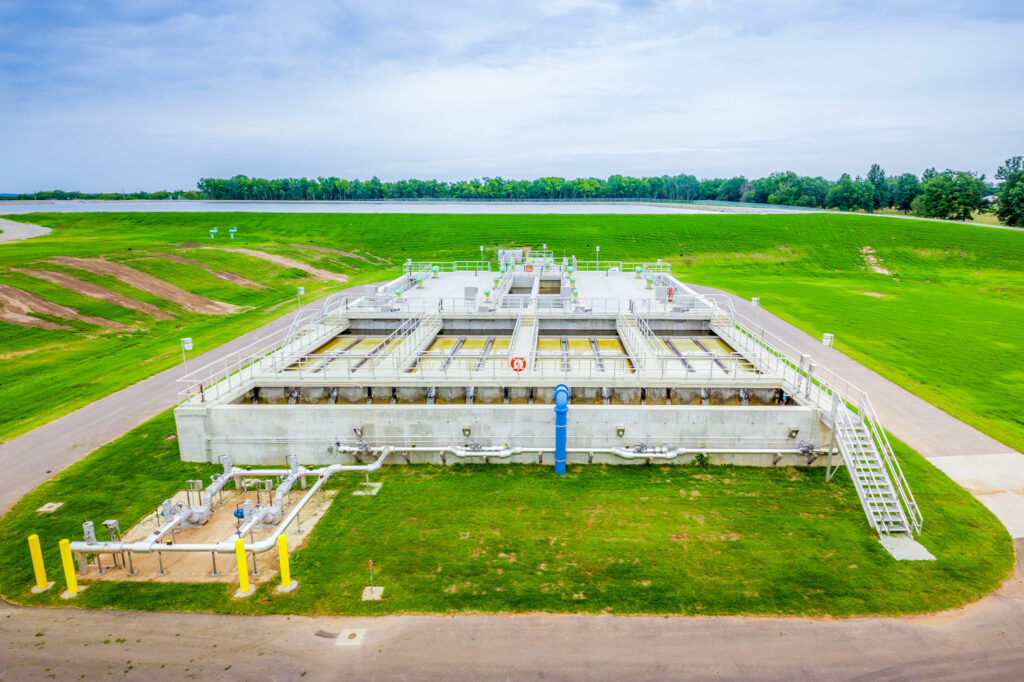 Aerial view of a modern water treatment plant surrounded by green grass, with walkways, equipment, and railings visible under a partly cloudy sky.