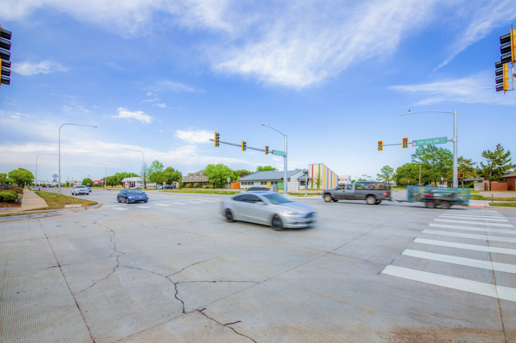 Several cars drive through a wide intersection with crosswalks, traffic signals, and a few buildings and trees in the background under a blue, partly cloudy sky.