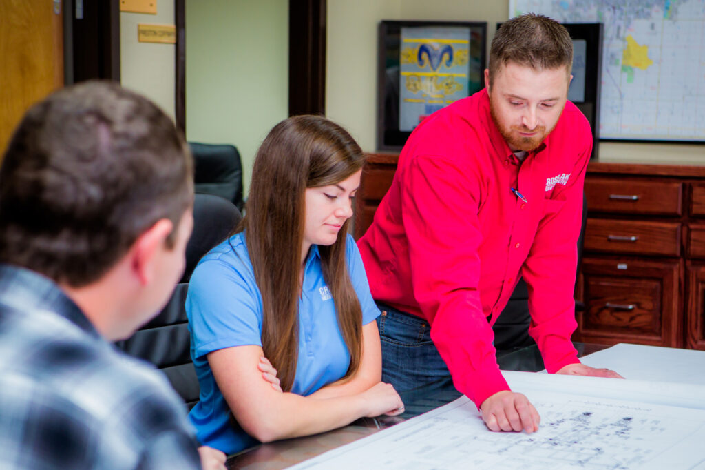 Three people are gathered around a table looking at large architectural blueprints, with one man in a red shirt pointing at the plans as he explains something to his colleagues.