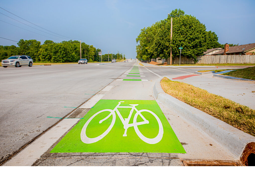 A bright green bike lane with a white bicycle symbol runs alongside a wide road, bordered by grass and trees, with cars driving in the adjacent lanes under a clear sky.