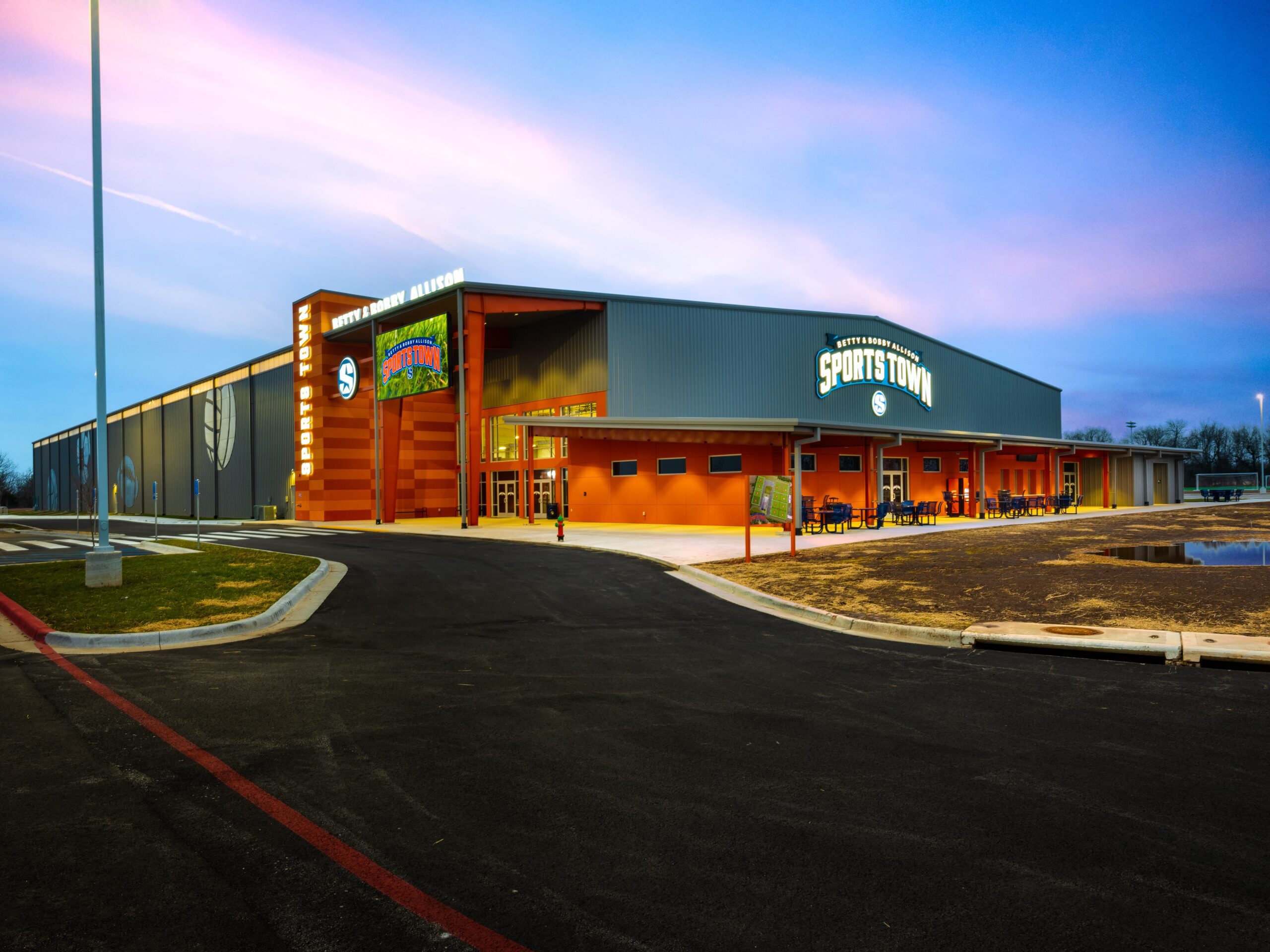 A modern sports complex with orange and gray exterior, large windows, and a "Sports Town" sign, surrounded by a parking lot at sunset. The sky is pink and blue, and lights illuminate the building.