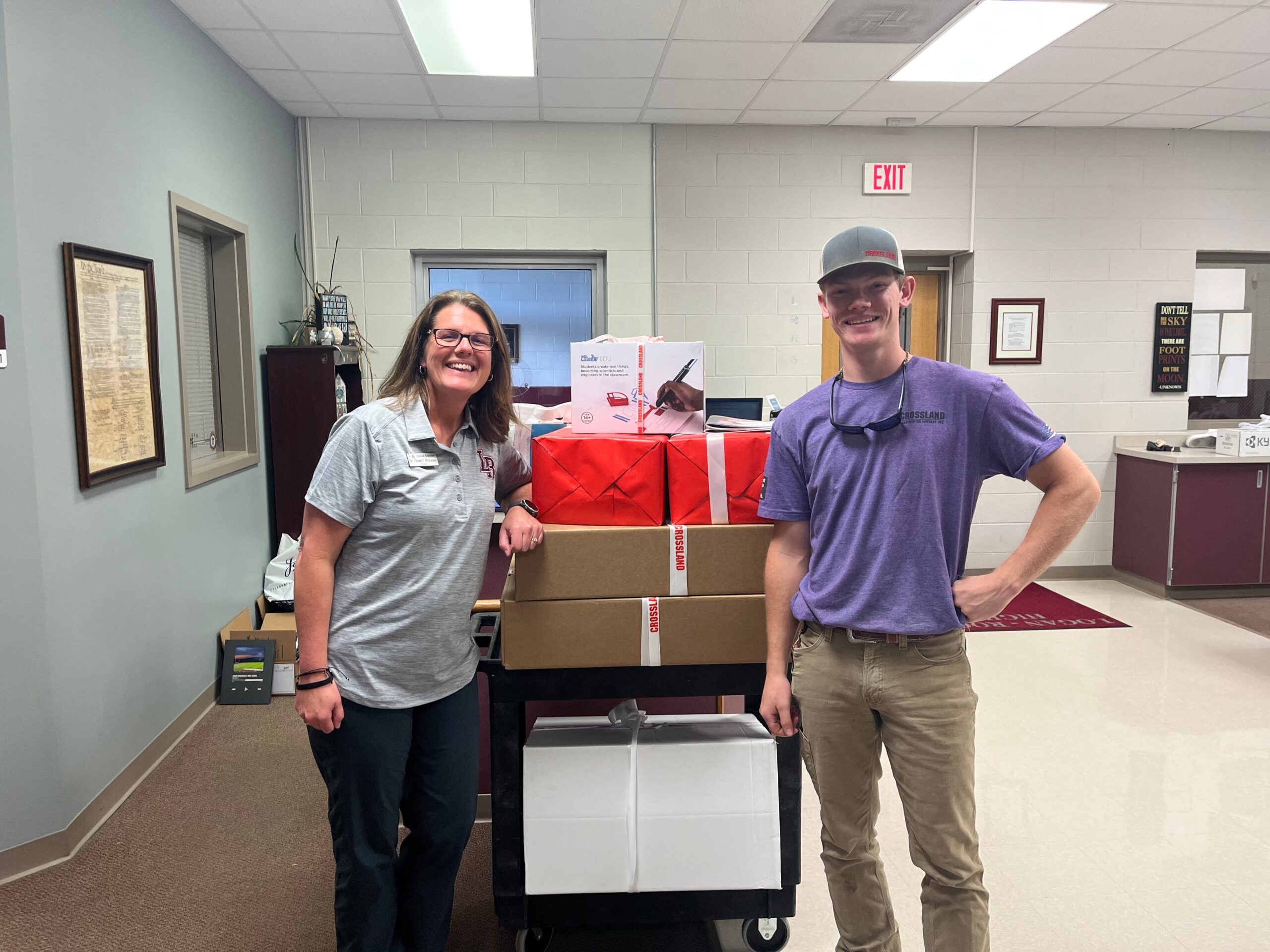 Two people stand smiling beside a cart stacked with large boxes and packages in an office setting. Fluorescent lights, tile floor, and framed documents on the wall are visible in the background.