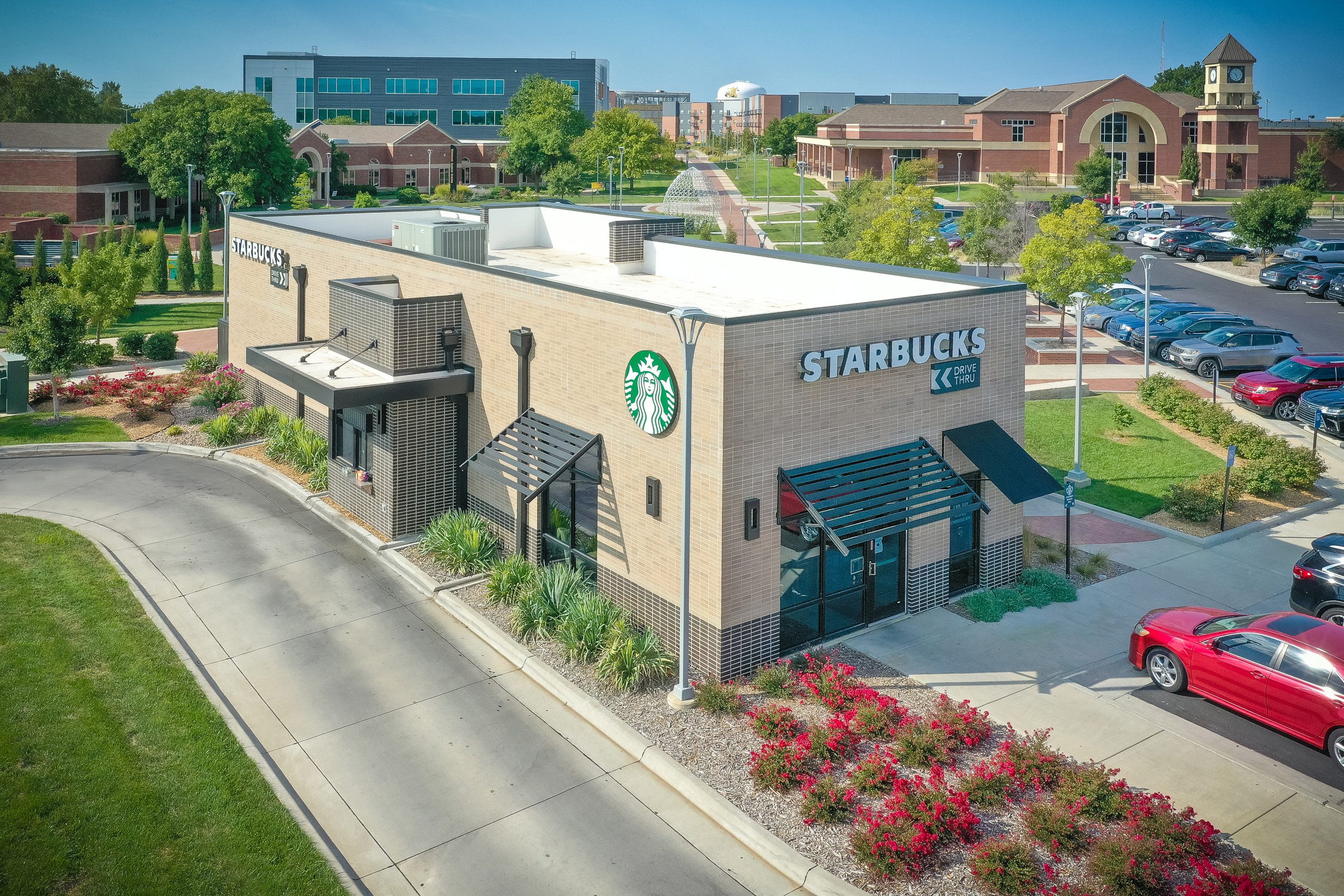 A modern standalone Starbucks building with drive-thru, surrounded by landscaping and parking, situated on a college campus with academic buildings in the background.