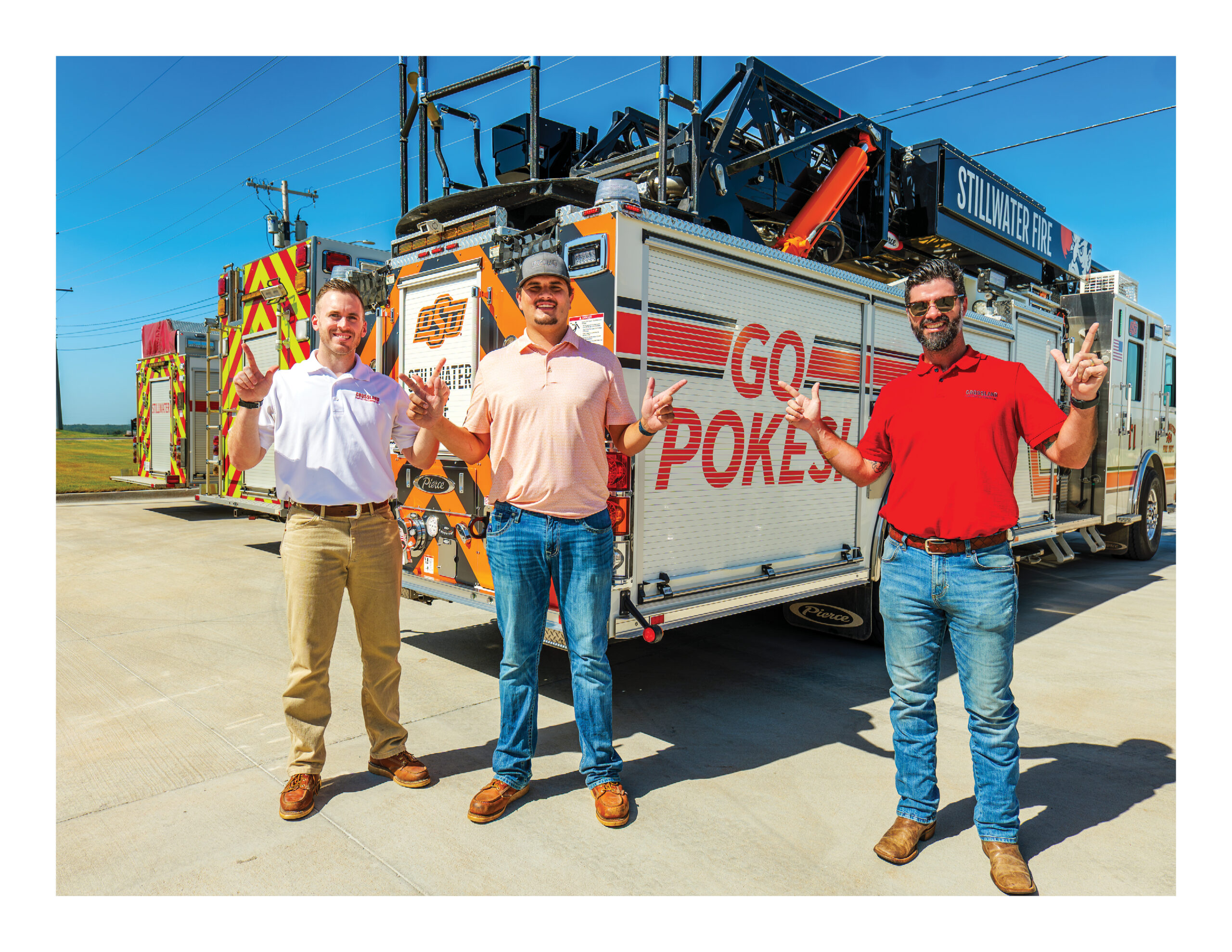 Three men stand in front of a fire truck that says “GO POKES!” on the side. They are outdoors on a sunny day, smiling, and making a hand gesture with two raised fingers.