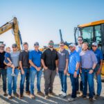 A group of eleven men and a young child stand together outdoors in front of large construction vehicles, wearing casual shirts, jeans, hats, and sunglasses, smiling on a sunny day at a construction site.