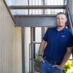 A man wearing a navy blue polo shirt and jeans leans casually against a metal railing under an outdoor staircase, smiling at the camera. Sunlight illuminates the scene and there is greenery in the background.