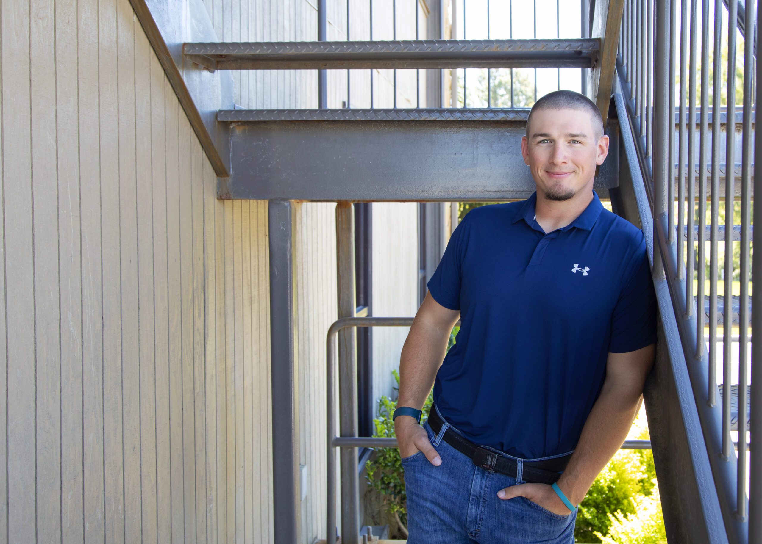 A man wearing a navy blue polo shirt and jeans leans casually against a metal railing under an outdoor staircase, smiling at the camera. Sunlight illuminates the scene and there is greenery in the background.