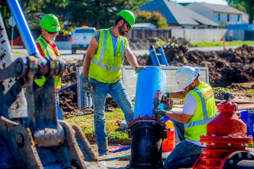 Three construction workers in safety vests and helmets work outdoors on installing or repairing a large blue pipe, surrounded by machinery, dirt, and equipment on a sunny day.