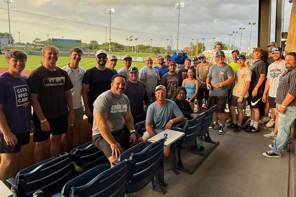 A group of people pose together in stadium seats at a baseball field during sunset, all smiling. Some wear hats and sunglasses. Stadium lights and flags are visible in the background.
