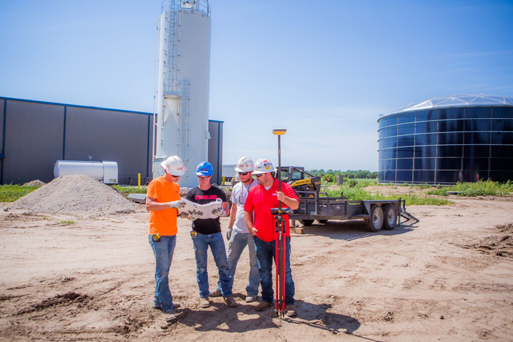 Four construction workers in hard hats stand on a construction site, reviewing plans. Behind them are a trailer, a large silo, and a black water tank, with clear blue sky overhead.