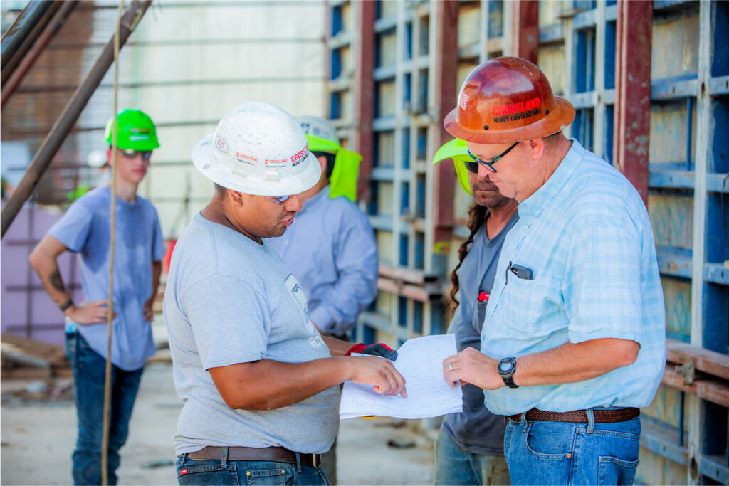 Two construction workers in hard hats review blueprints at a job site, while others in safety gear stand nearby. Scaffolding and building materials surround them, indicating active construction work.