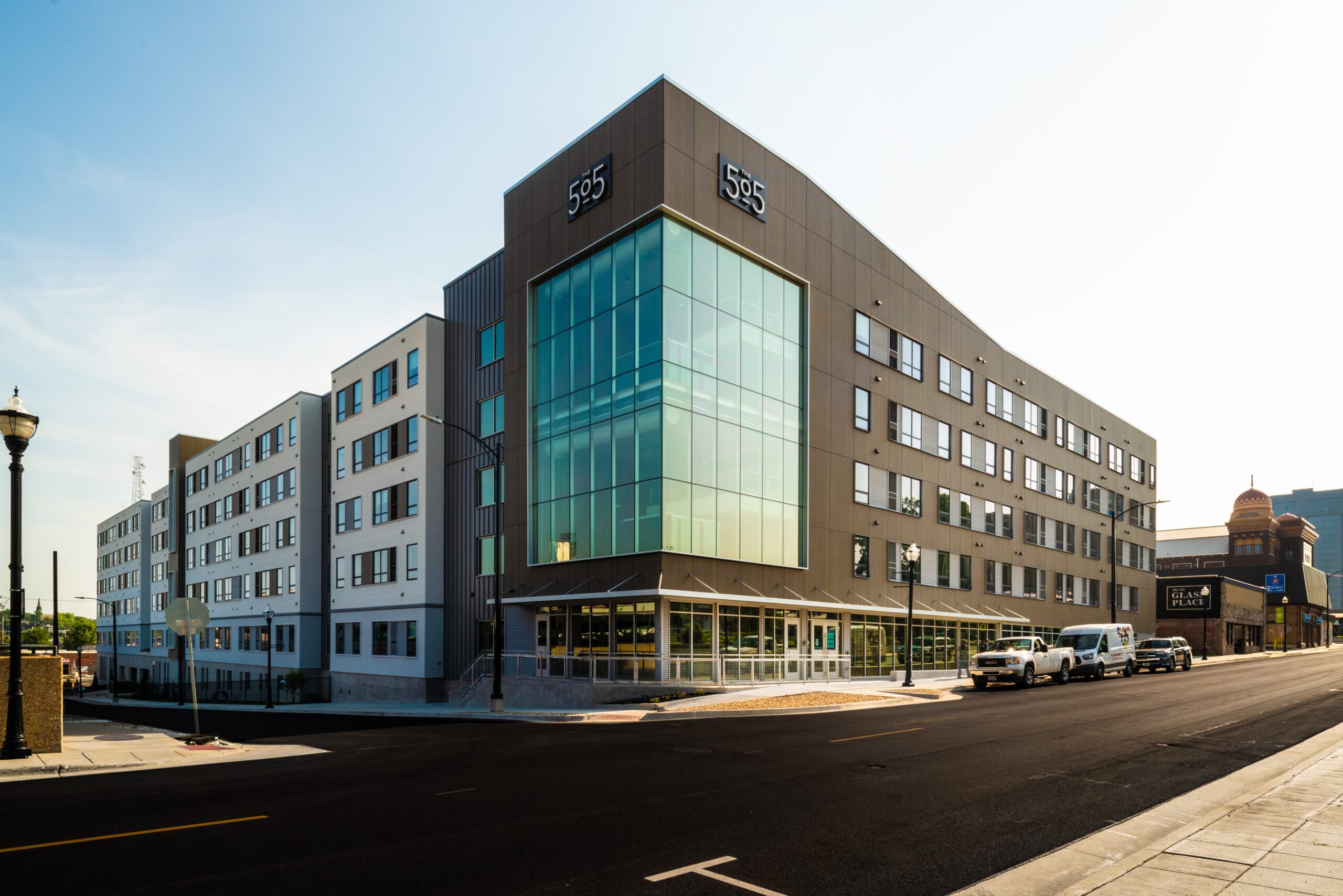 A modern, multi-story apartment building with large glass windows on the corner and “555” displayed on its facade, located on a clean, empty street with a few parked cars and clear sky above.