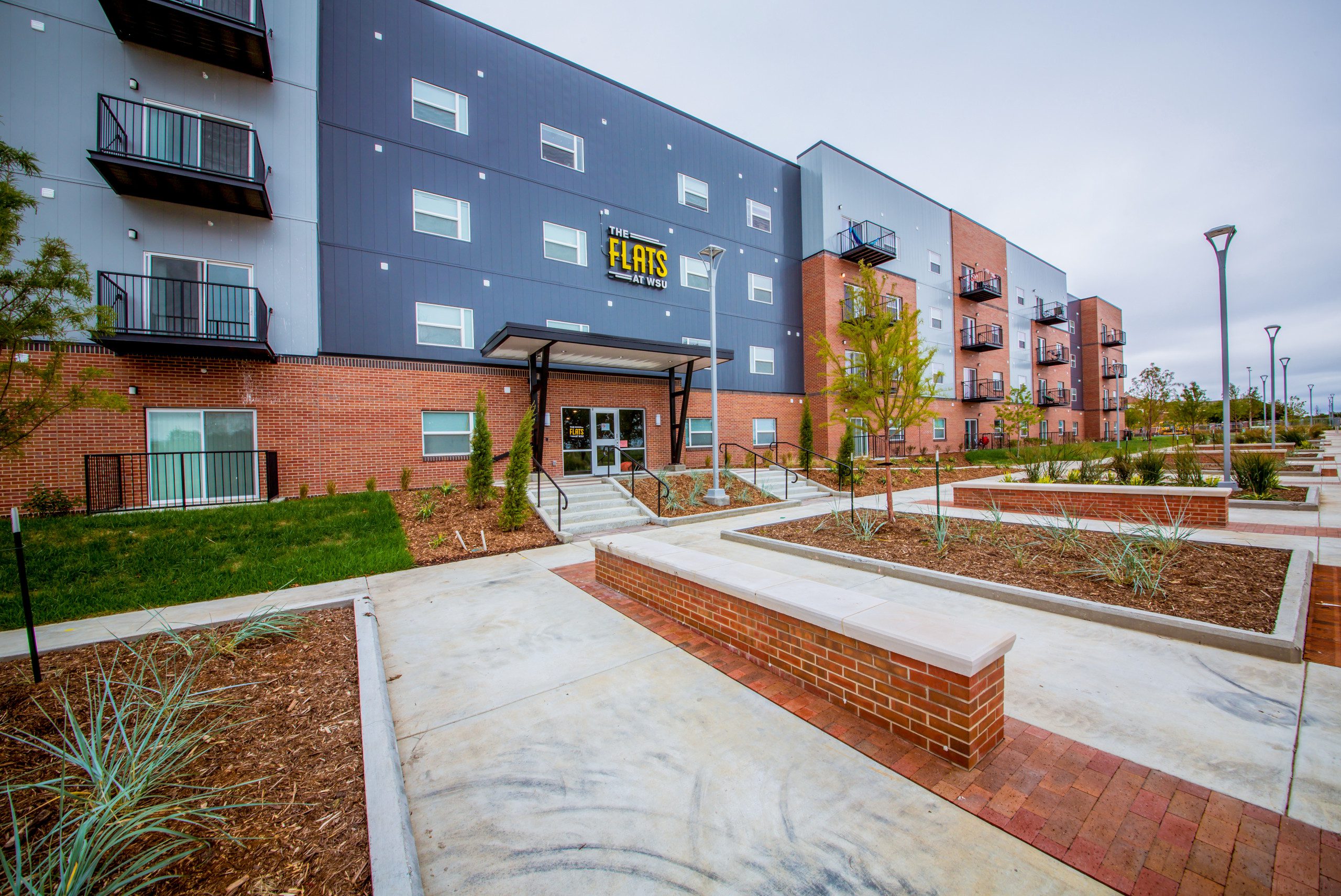 Modern apartment building with three stories, featuring balconies, a sign reading "FLATS," brick and gray panel exterior, landscaped pathways, young trees, and benches in front of the entrance.