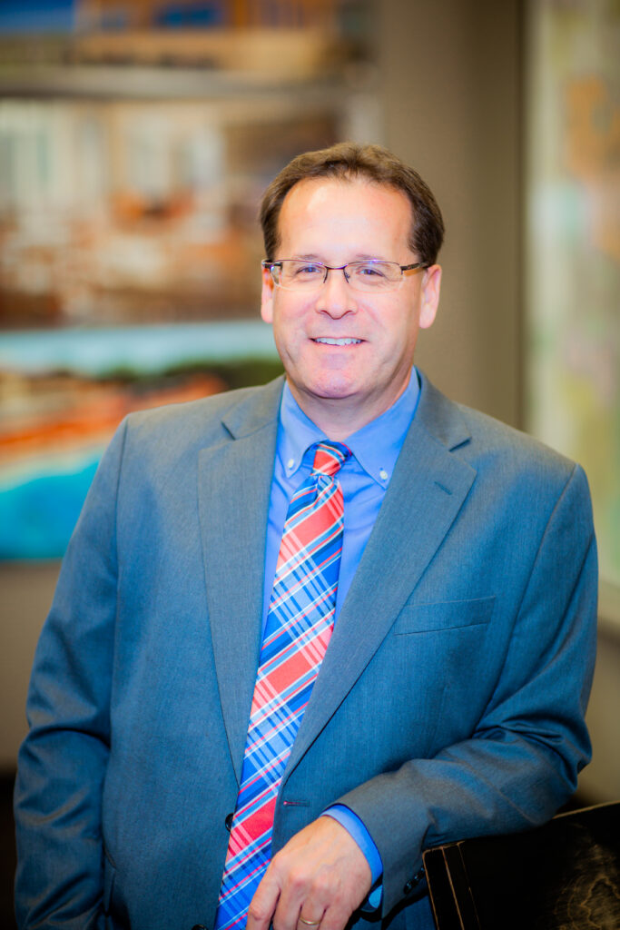 A man in a blue suit and colorful plaid tie stands indoors, smiling and leaning on a surface, with blurred office artwork in the background.