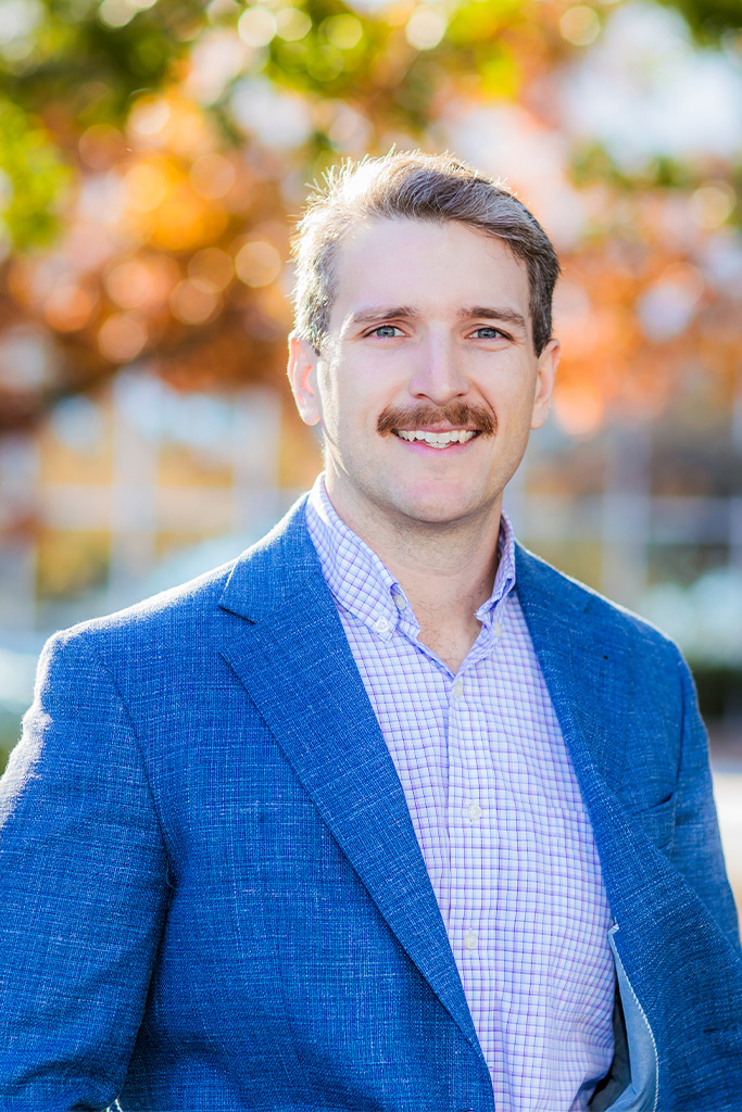 A man with light brown hair and a mustache, wearing a blue blazer and checked shirt, smiles outdoors with blurred autumn foliage in the background.
