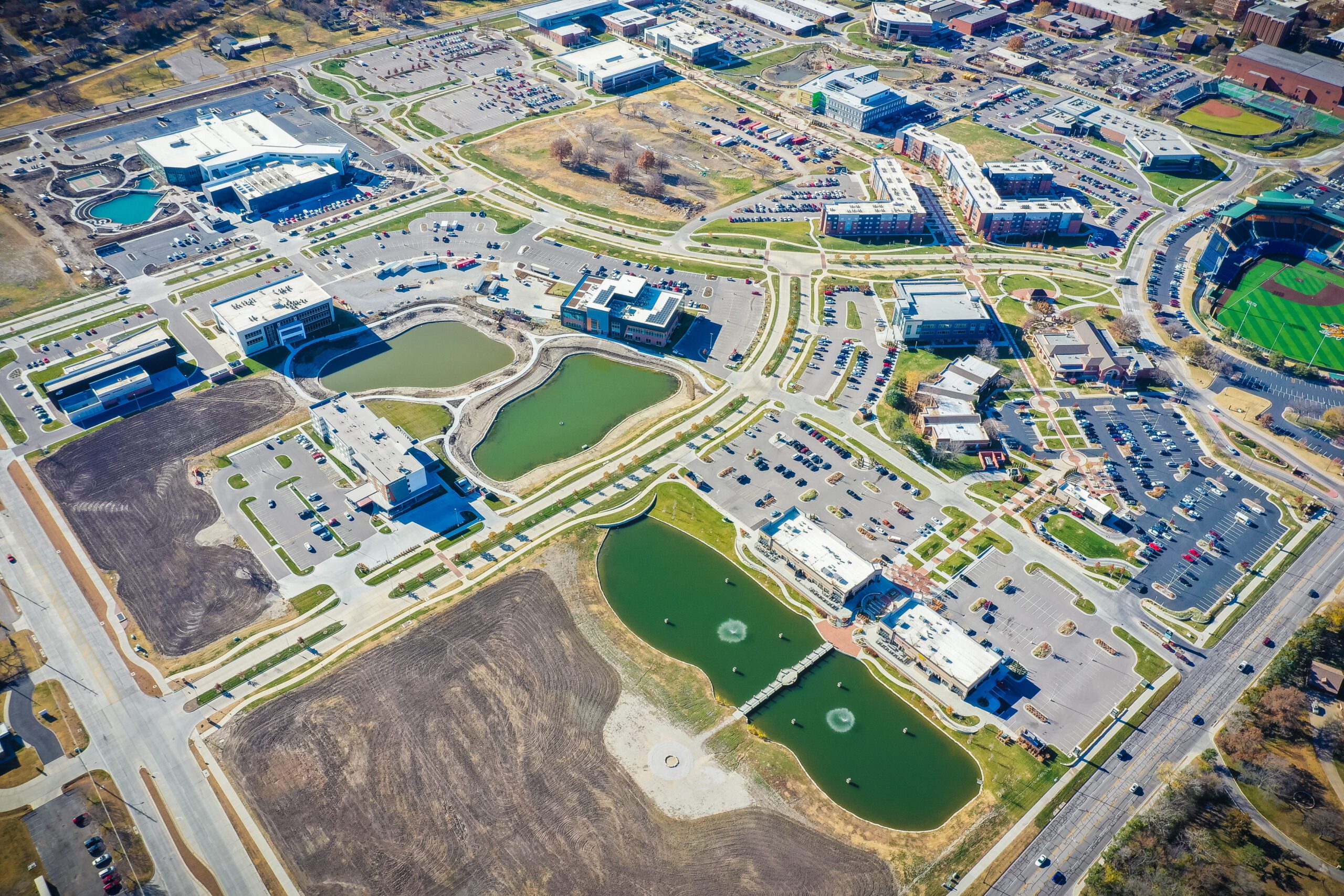 Aerial view of a suburban area with office buildings, parking lots, green spaces, ponds with fountains, sports fields, and roads connecting the properties.