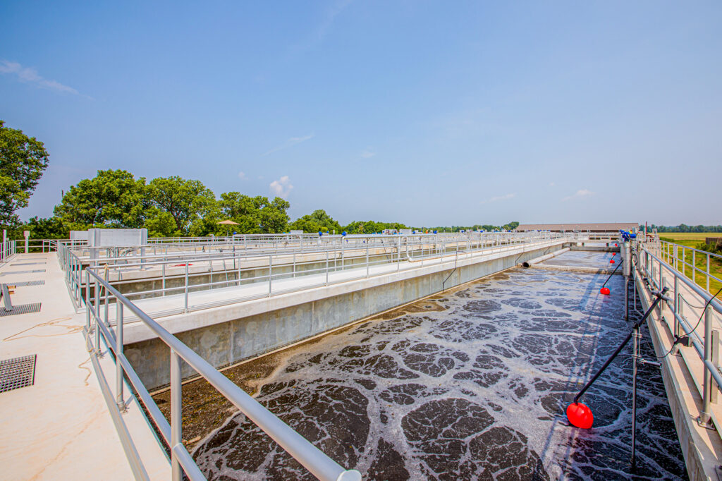 A modern outdoor wastewater treatment facility with concrete tanks, metal railings, and aerated water. Trees and grass surround the plant under a clear blue sky.