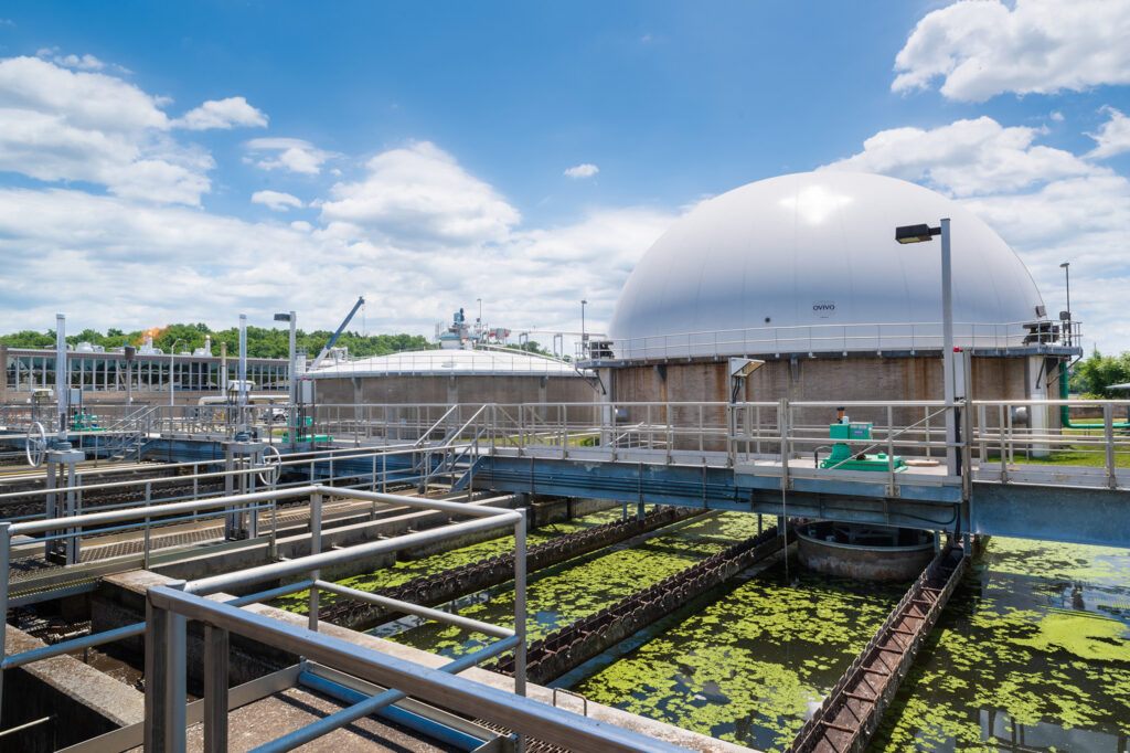 Industrial water treatment plant with large domed tanks, metal walkways, and water basins partially covered in green algae, under a bright blue sky with scattered clouds.