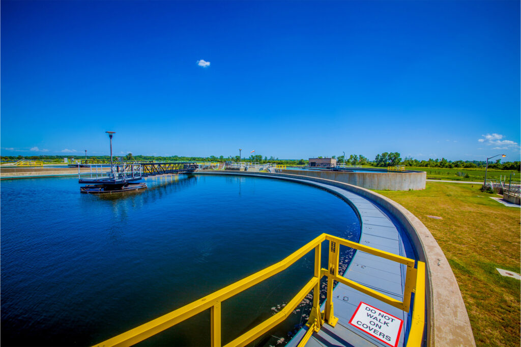 A large circular water treatment basin with clear blue water, surrounded by a yellow railing and green grass under a bright blue sky. A sign reads, “DO NOT WALK ON COVERS.”.
