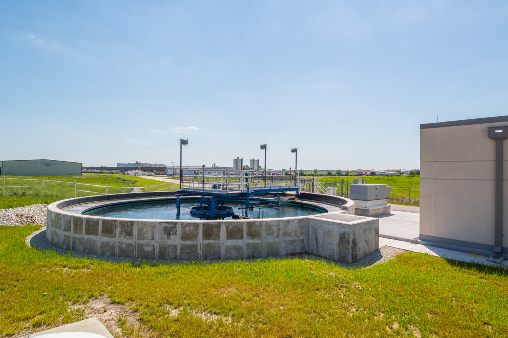 A large circular water treatment tank sits outdoors on a concrete base, surrounded by grass, with industrial buildings in the background under a clear blue sky.