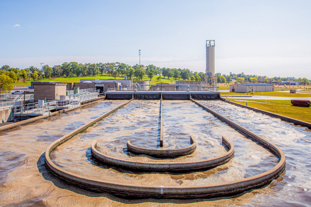 A wastewater treatment plant with circular settling tanks, aeration basins, and industrial buildings, surrounded by green grass and trees under a clear sky.