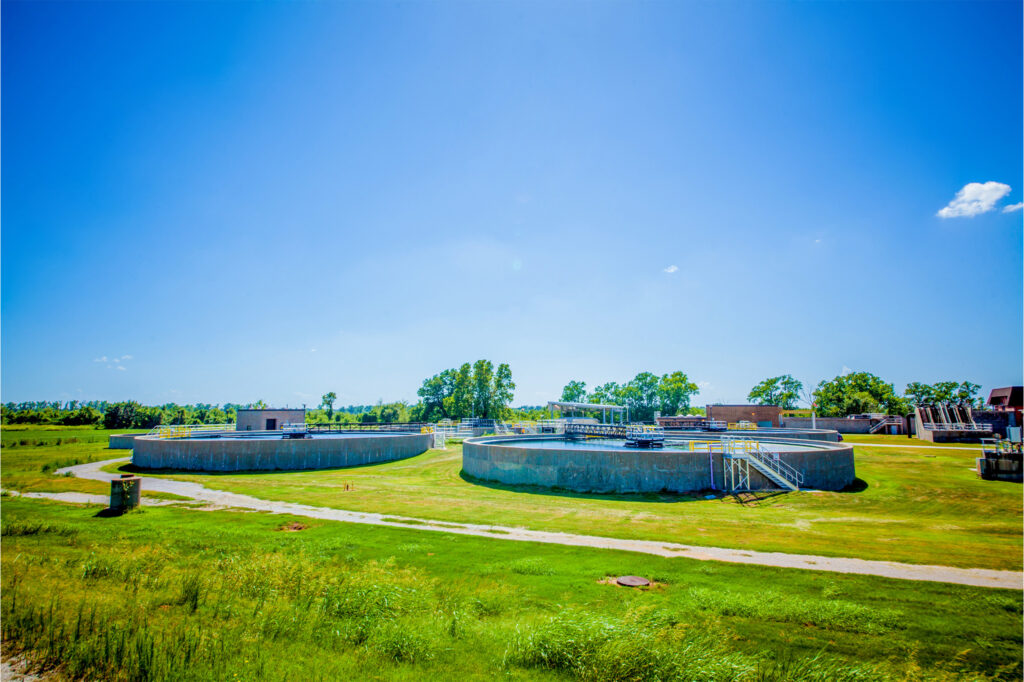 A wastewater treatment plant with large circular tanks, surrounded by green grass and trees under a bright blue sky on a sunny day.