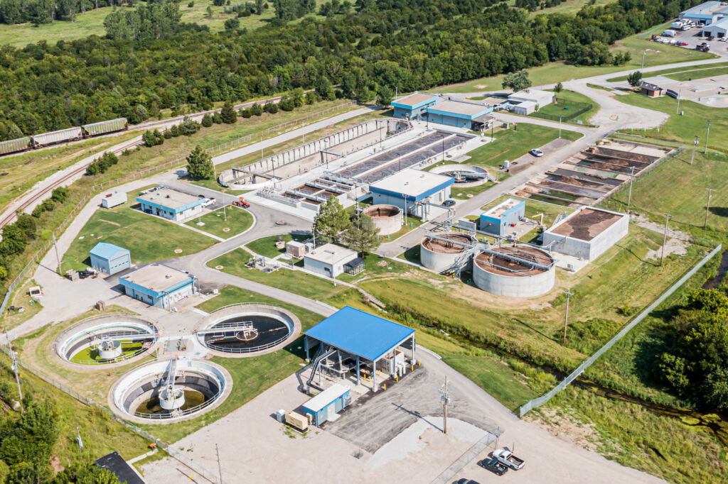 Aerial view of a water treatment plant with circular and rectangular tanks, blue-roofed buildings, surrounded by grassy fields and trees, with roads and railway nearby.