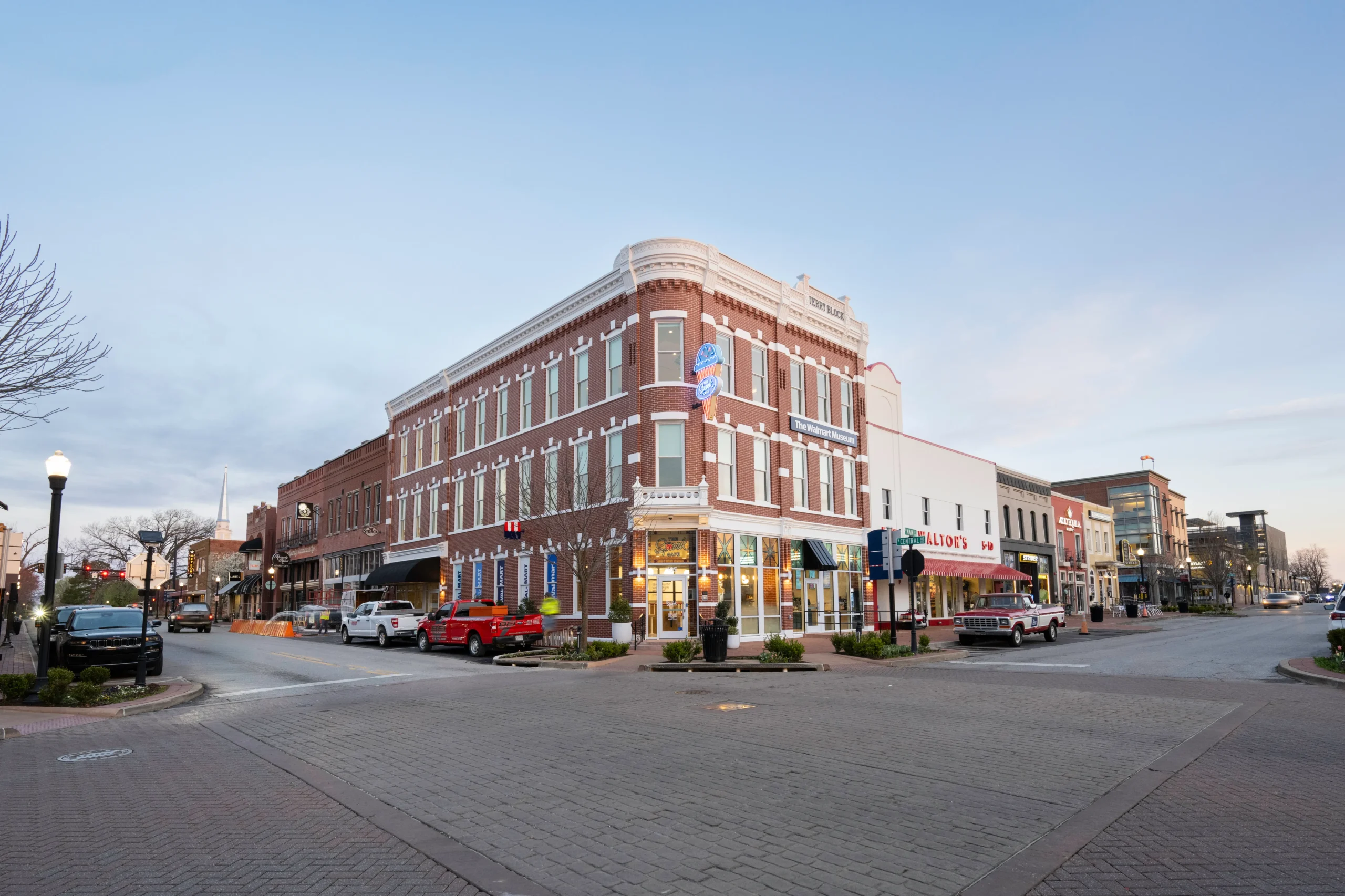 A historic downtown street corner features a large red-brick building with shops on the ground floor, parked cars along the street, and evening lighting under a clear sky.