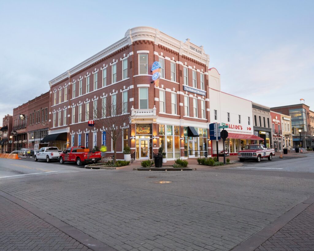 A red brick corner building with white trim houses shops and restaurants on a quiet street, with several parked trucks and a few small trees along the sidewalk. The sky is clear and buildings line both sides of the street.