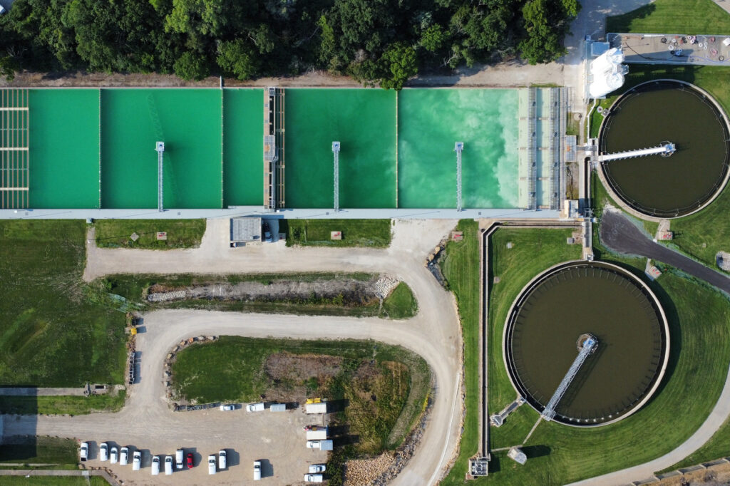 Aerial view of a water treatment plant showing rectangular green water tanks, two large circular tanks, surrounding roads, parked vehicles, and trees at the top edge of the image.
