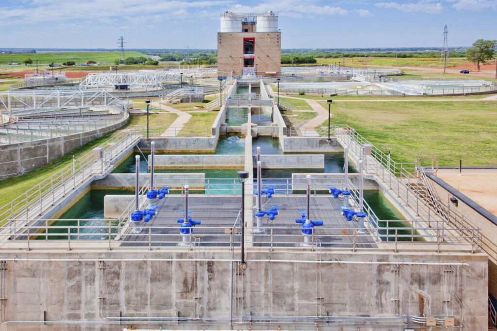 A water treatment plant with large tanks, pipes, and machinery, surrounded by grass and open fields under a blue sky with some clouds.