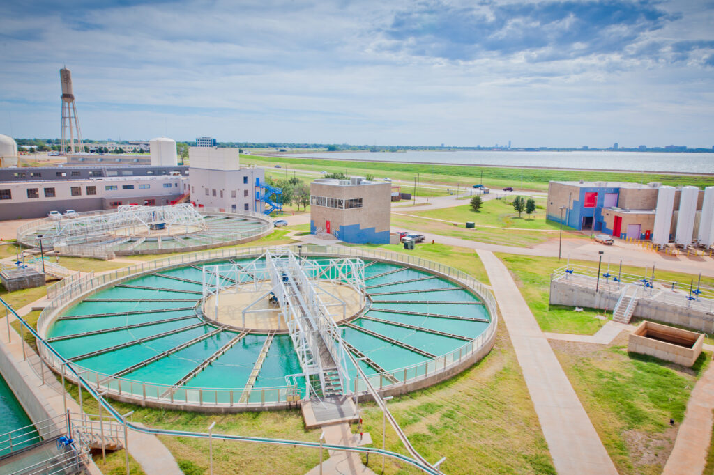 A water treatment plant with large circular tanks, buildings, and walkways on a grassy area near a reservoir, under a partly cloudy sky.