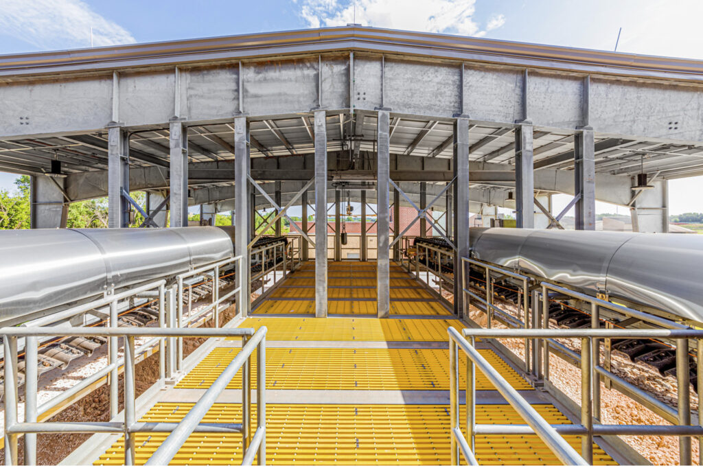 A modern, elevated pedestrian bridge with yellow grated flooring and metal railings spans over train tracks, with steel beams and a roof overhead.