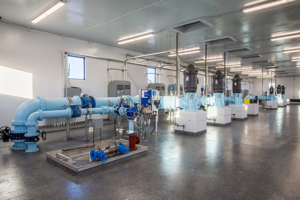 A clean, well-lit industrial room with large blue pipes, valves, and several electric pumps is visible. The space houses modern water supply or wastewater treatment equipment.