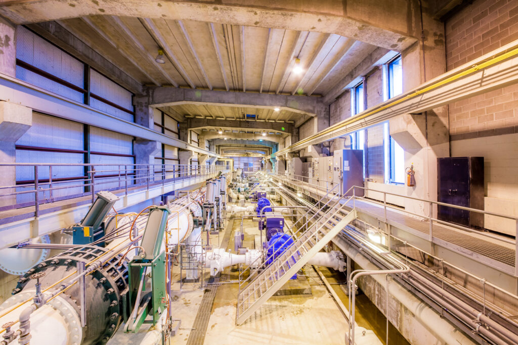 Industrial facility interior with large pipes, valves, and machinery, including blue pumps. Overhead lighting and concrete beams are visible, with metal walkways and railings running alongside the equipment.