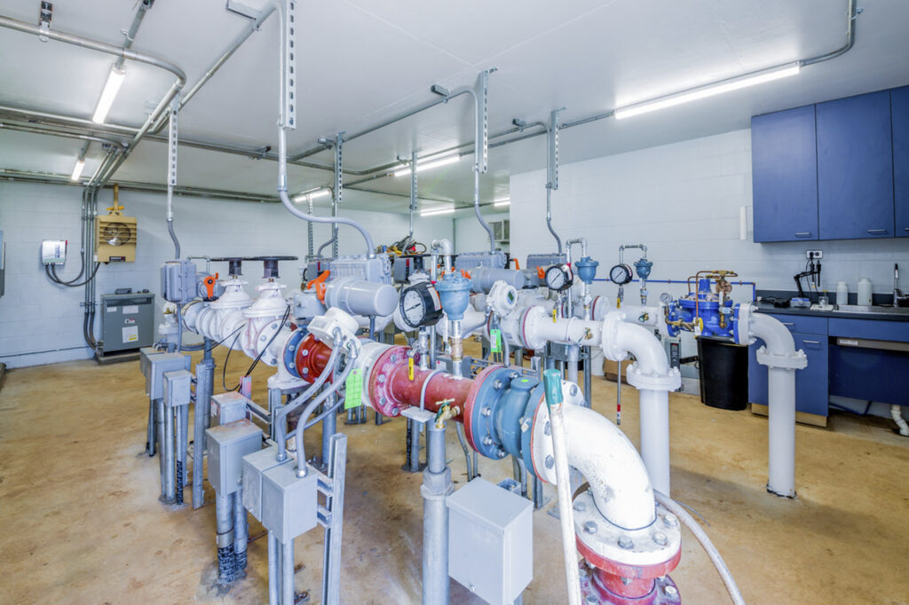 Interior view of a utility room with water pipes, gauges, control panels, and blue cabinets along the wall; the floor is tan, and the walls and ceiling are white.