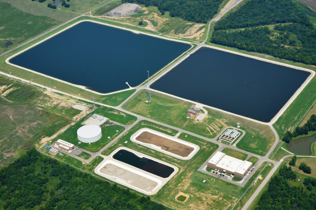 Aerial view of a water treatment facility with large rectangular pools, storage tanks, and several buildings surrounded by green fields and trees.