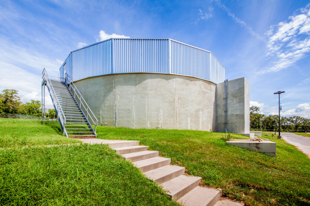 A large circular concrete water tank with a metal railing, an external metal staircase, and steps leading up a grassy slope under a bright blue sky with scattered clouds.
