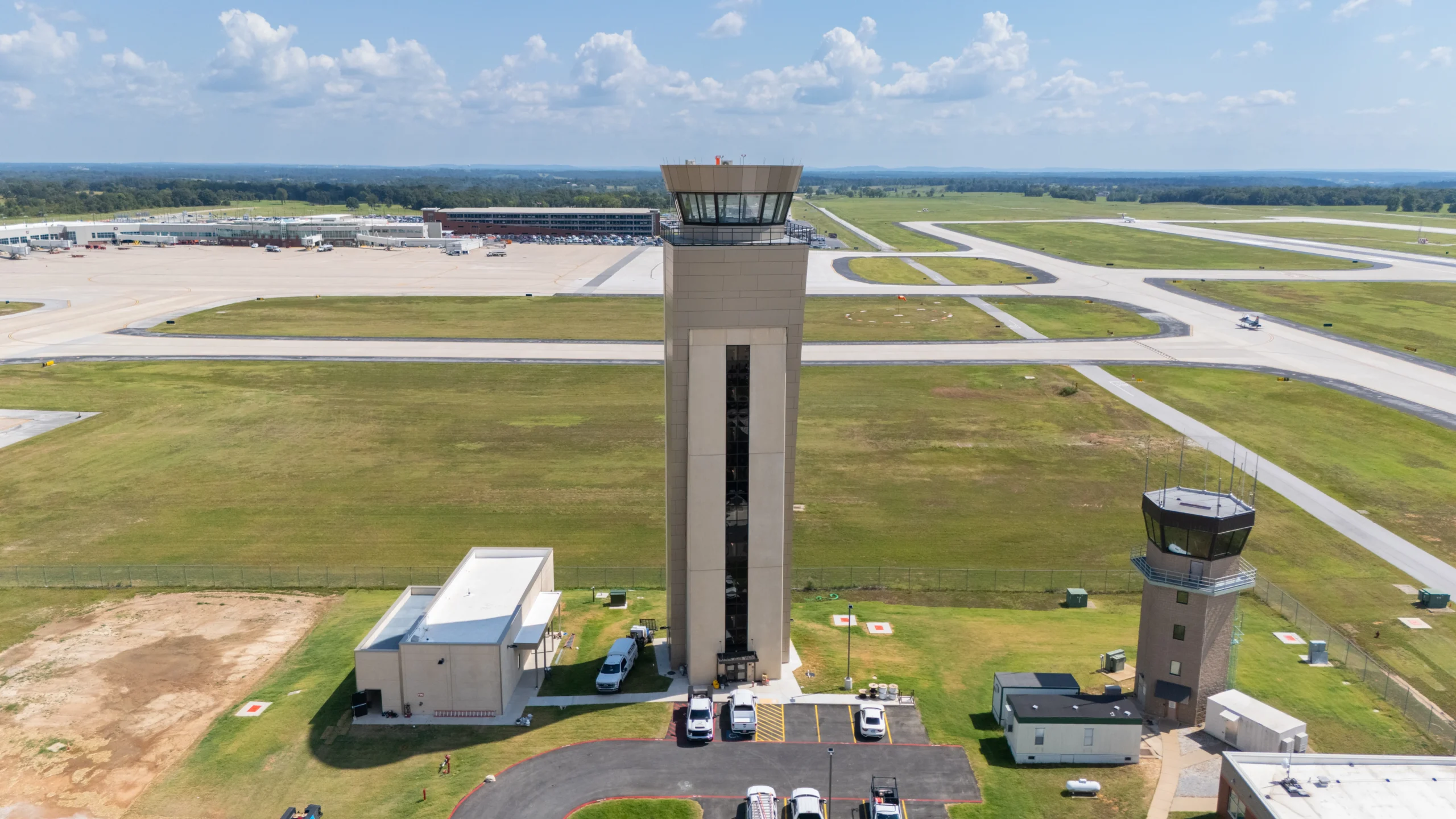 A tall airport control tower stands in the center of an airfield, surrounded by grassy areas, taxiways, and runways. Several smaller buildings and parked vehicles are visible at the base of the tower.