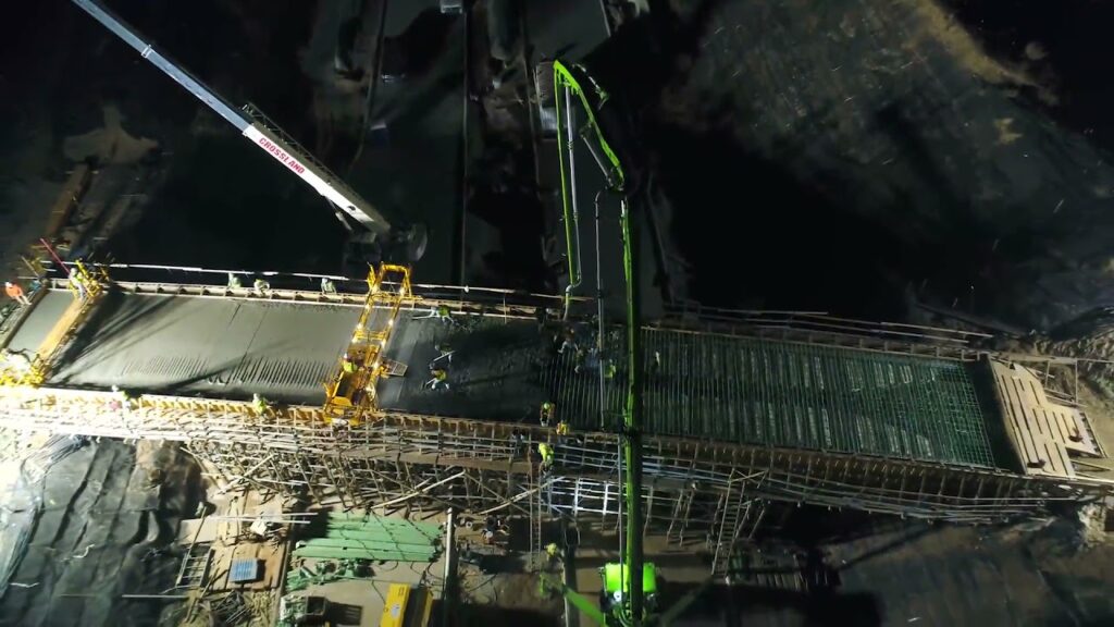 Aerial view of a nighttime construction site, showing workers and machinery pouring concrete and building the framework of a large bridge or elevated structure under bright artificial lighting.