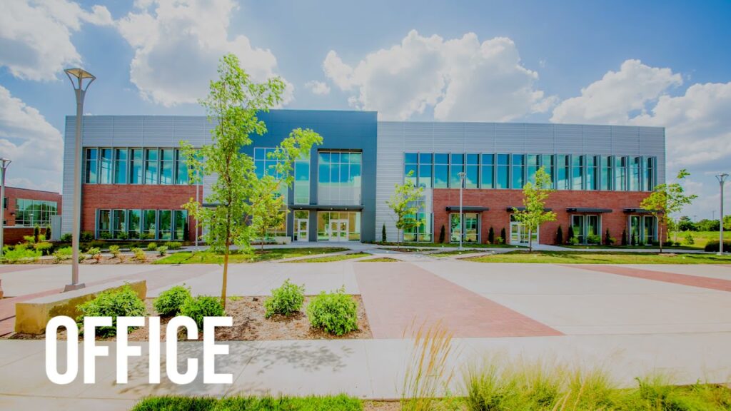 Modern two-story office building with large windows, surrounded by landscaped greenery and trees under a bright, partly cloudy sky. The word "OFFICE" is in bold white text at the bottom left.