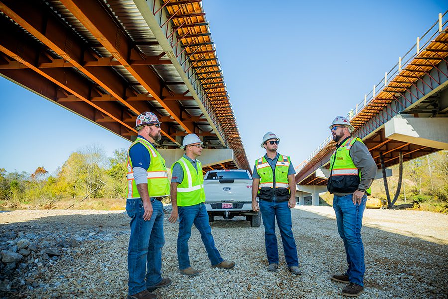 Four construction workers wearing safety vests and helmets stand and talk beneath a large, partially constructed bridge, with a white pickup truck parked nearby on a gravel surface.